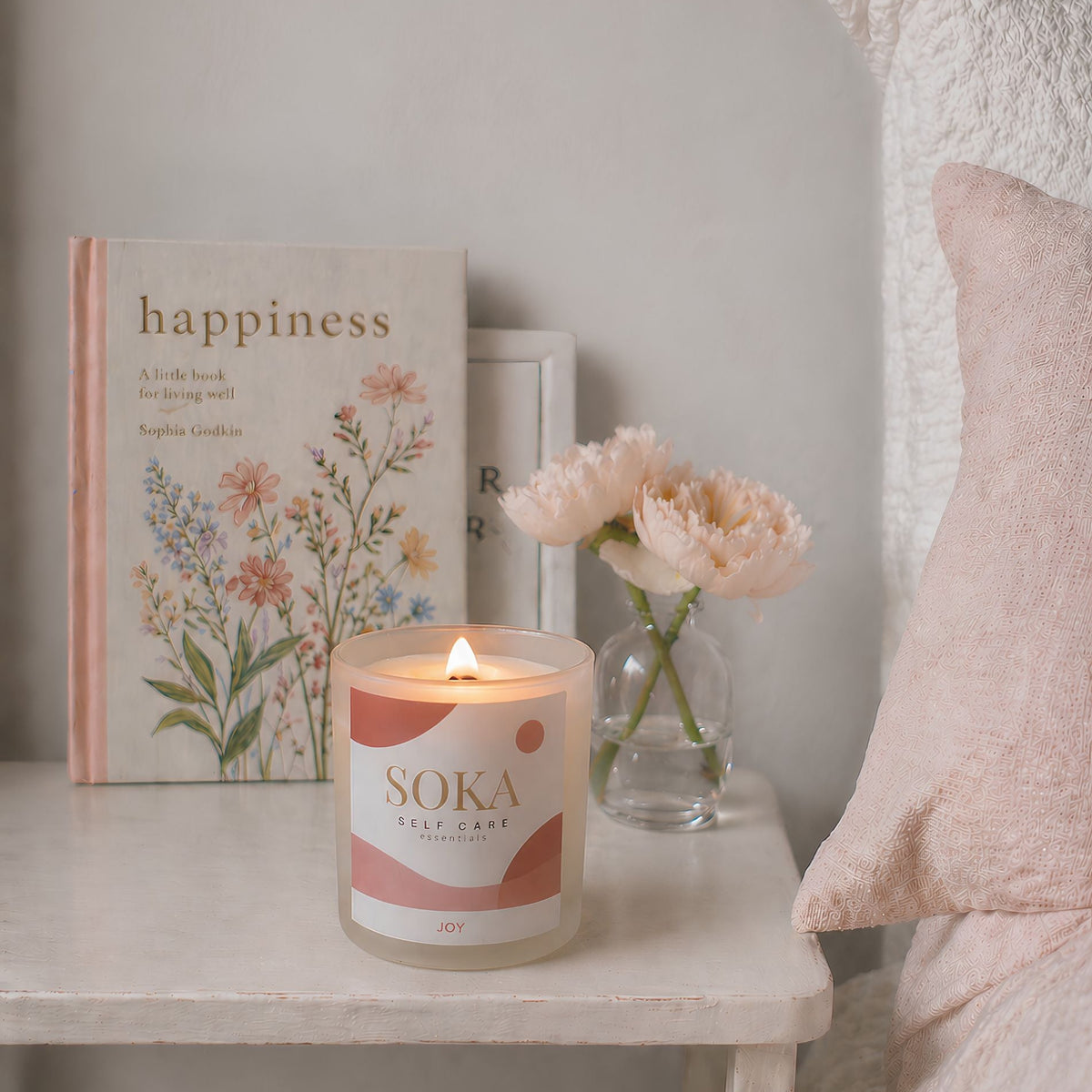 JOY Soka candle on a nightstand with a book titled 'happiness' and a vase of flowers in the background.