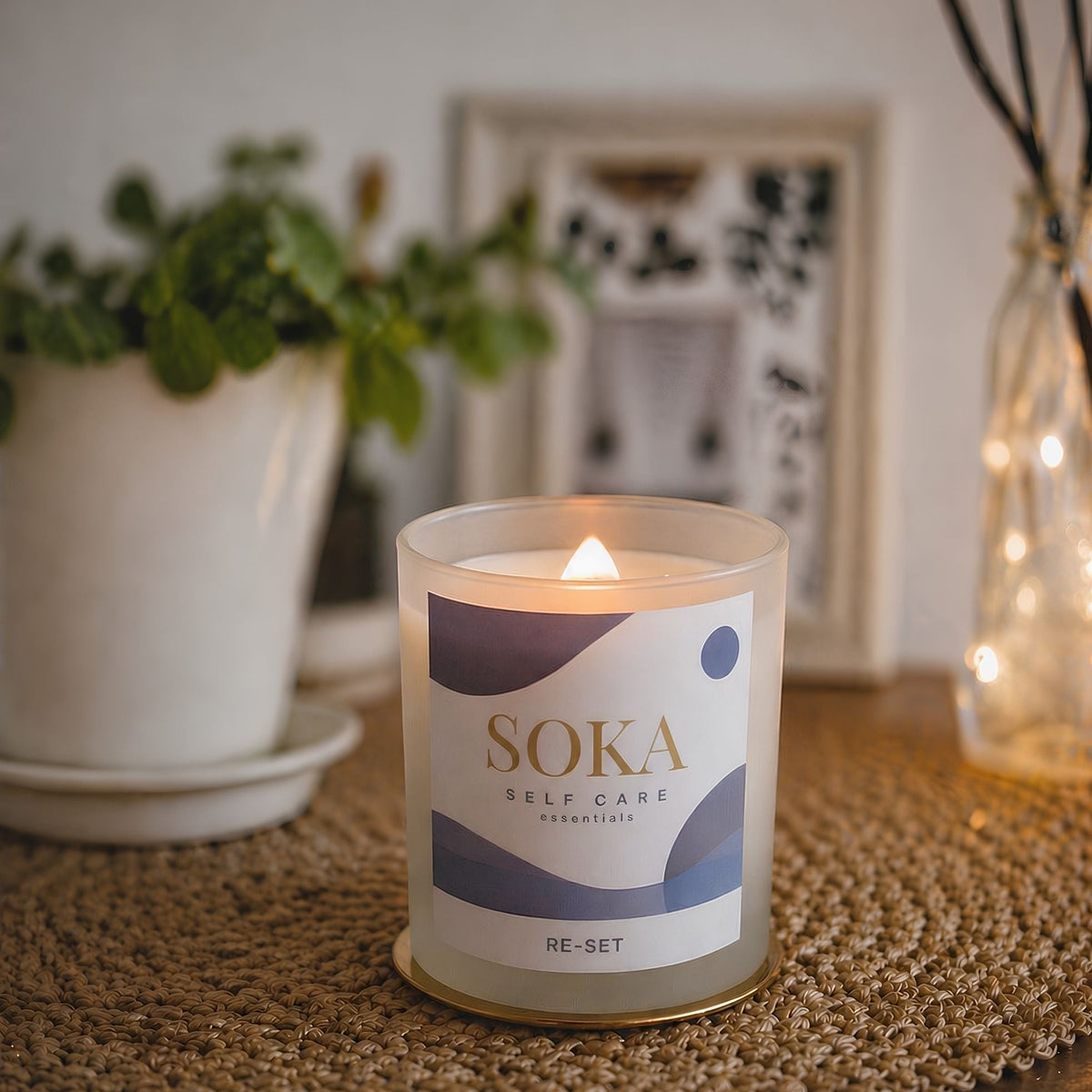 RE-SET Soka self-care candle on a textured surface with a plant and framed picture in the background